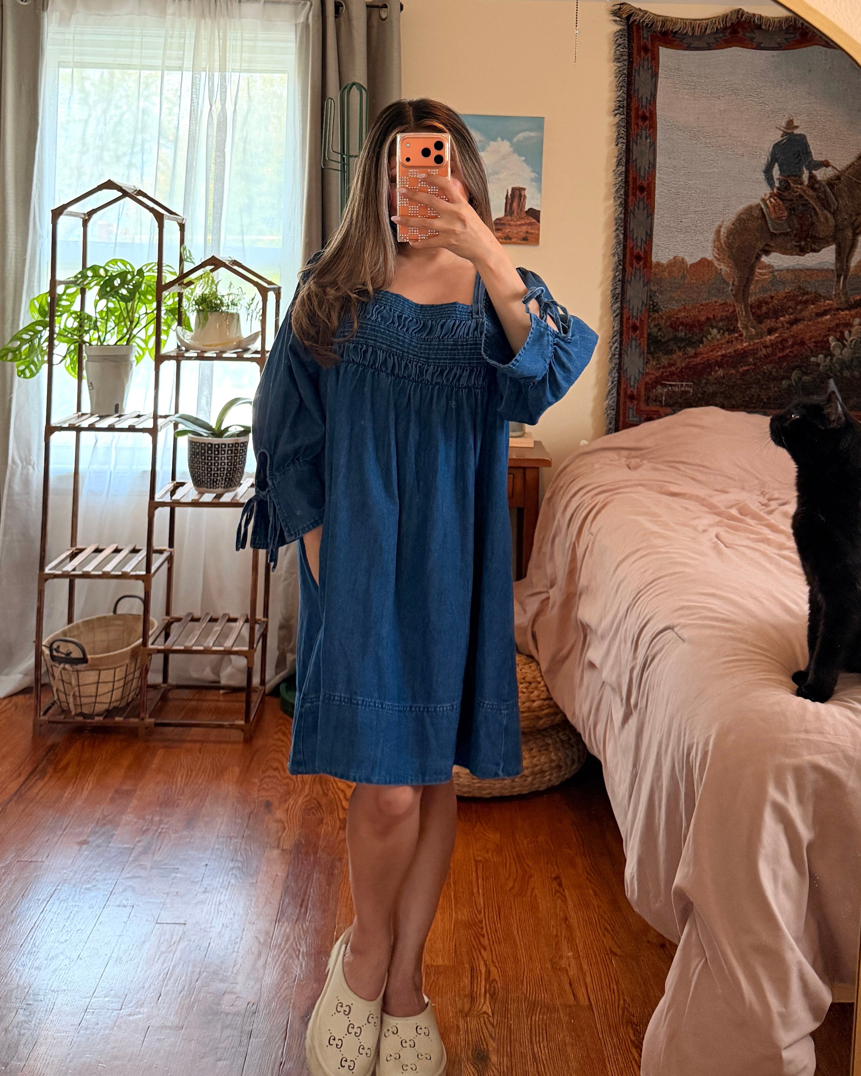 A woman takes a mirror selfie in a bedroom, wearing a blue smocked-embroidered long-sleeve dress with tie details at the cuffs. A black cat sits on the bed in the background, and a gold-framed archway surrounds the mirror.