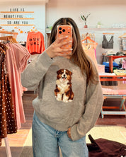 A woman taking a mirror selfie in a clothing boutique, wearing a gray crewneck sweatshirt with a large brown and white dog graphic on the front, paired with light blue jeans.