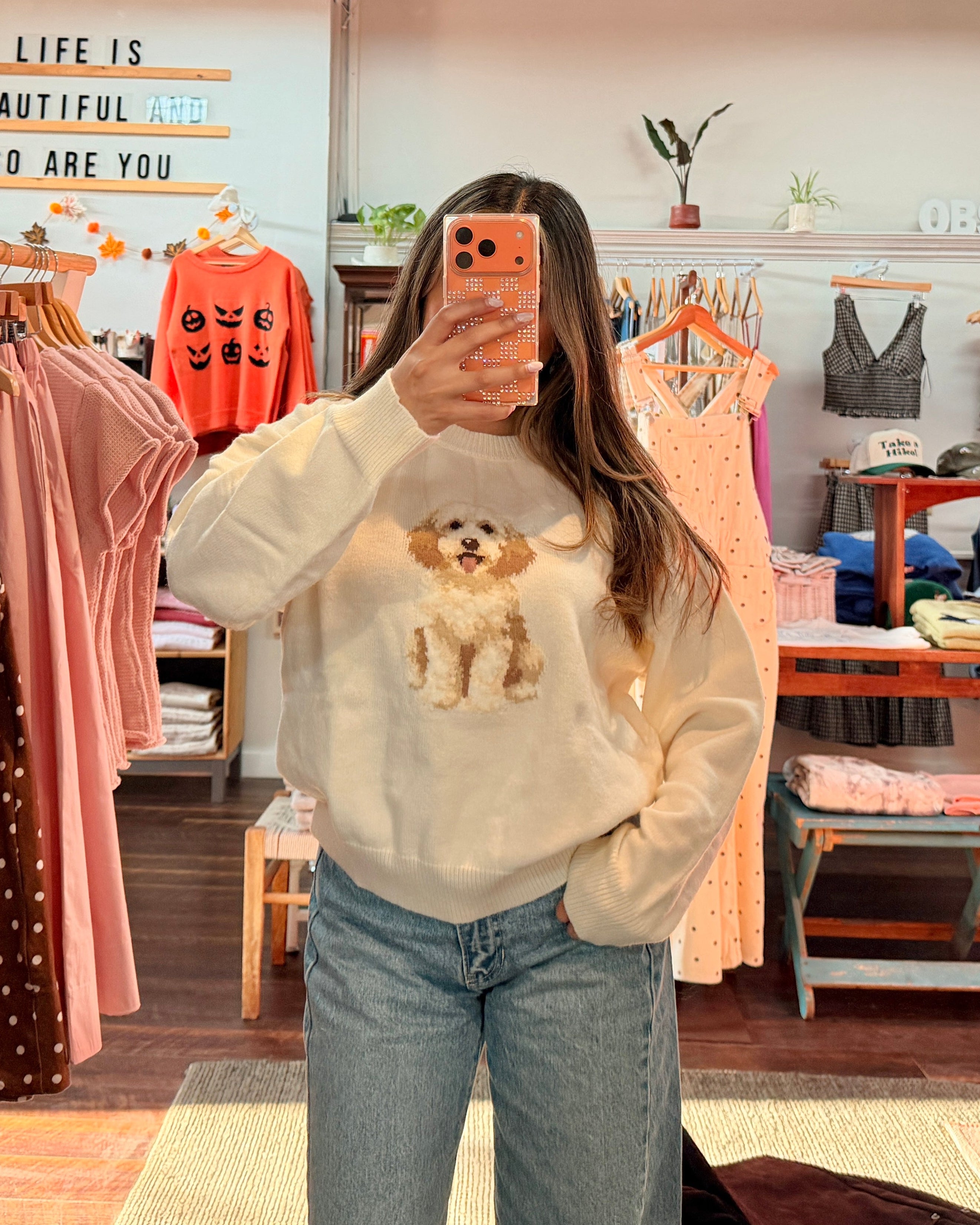 A woman takes a mirror selfie in a clothing boutique wearing a cream-colored crewneck sweatshirt with a dog graphic and light-wash blue jeans. The boutique is filled with racks of colorful clothing, wooden tables, and store fixtures.