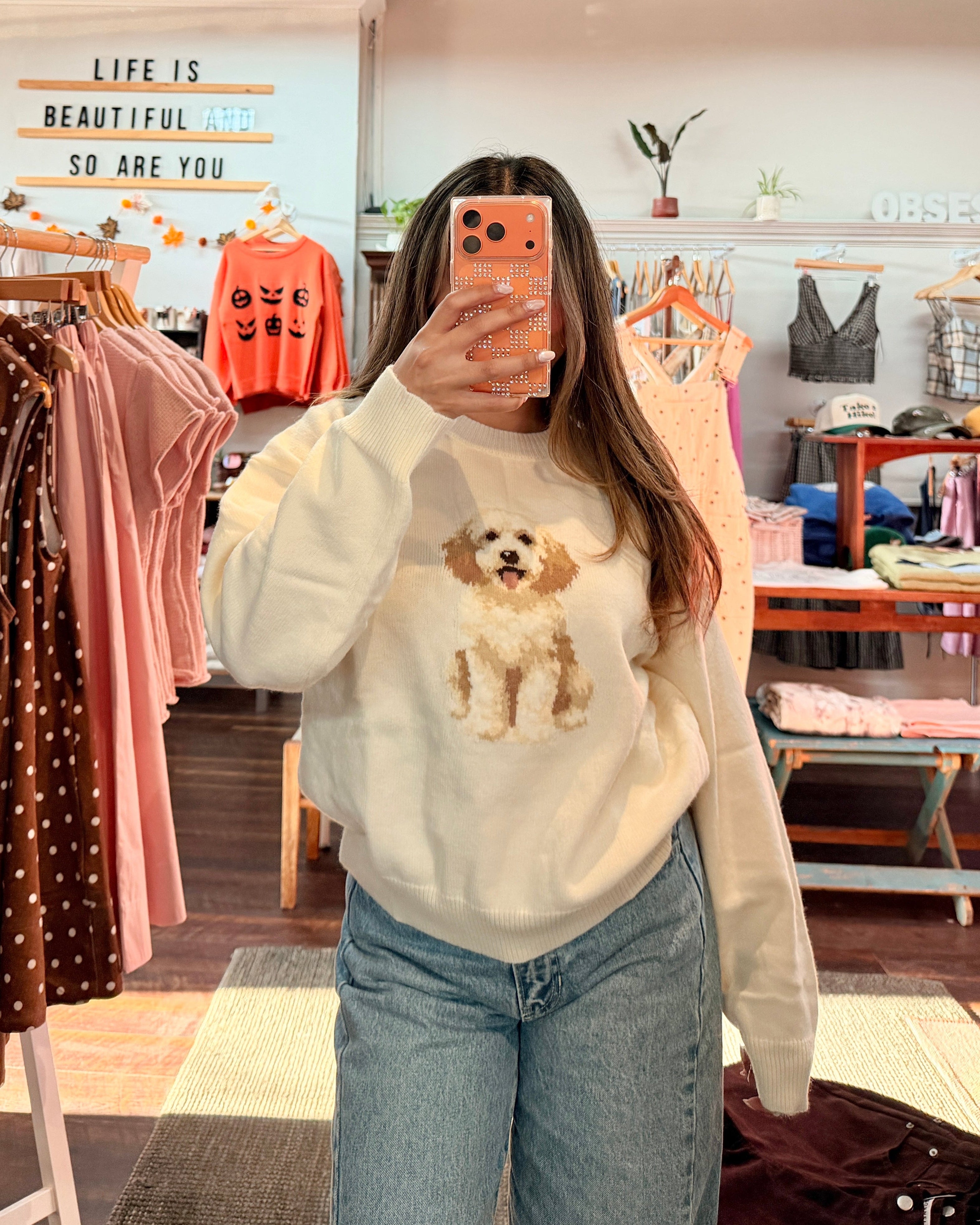 A woman takes a mirror selfie in a clothing boutique wearing a cream-colored sweatshirt with a dog graphic and light blue jeans. Clothing racks and store fixtures are visible in the background.