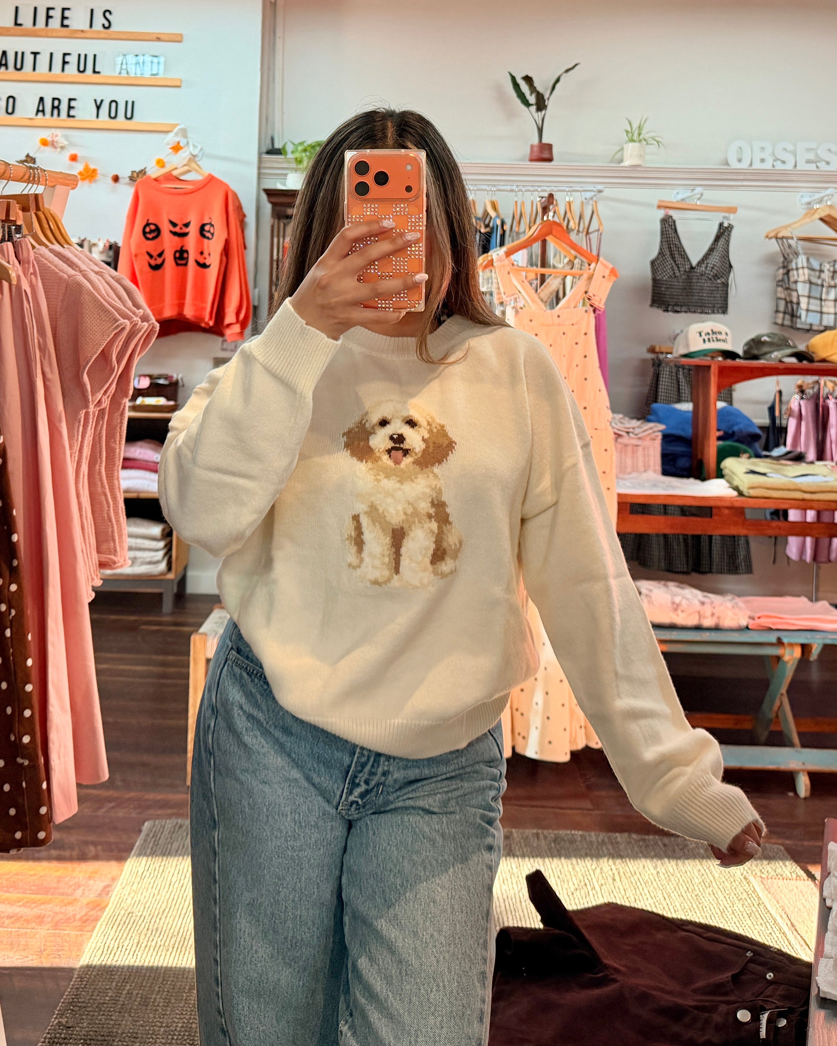 A woman takes a mirror selfie in a clothing boutique wearing a cream-colored long-sleeve sweater with a dog graphic on the front and light-wash blue jeans.
