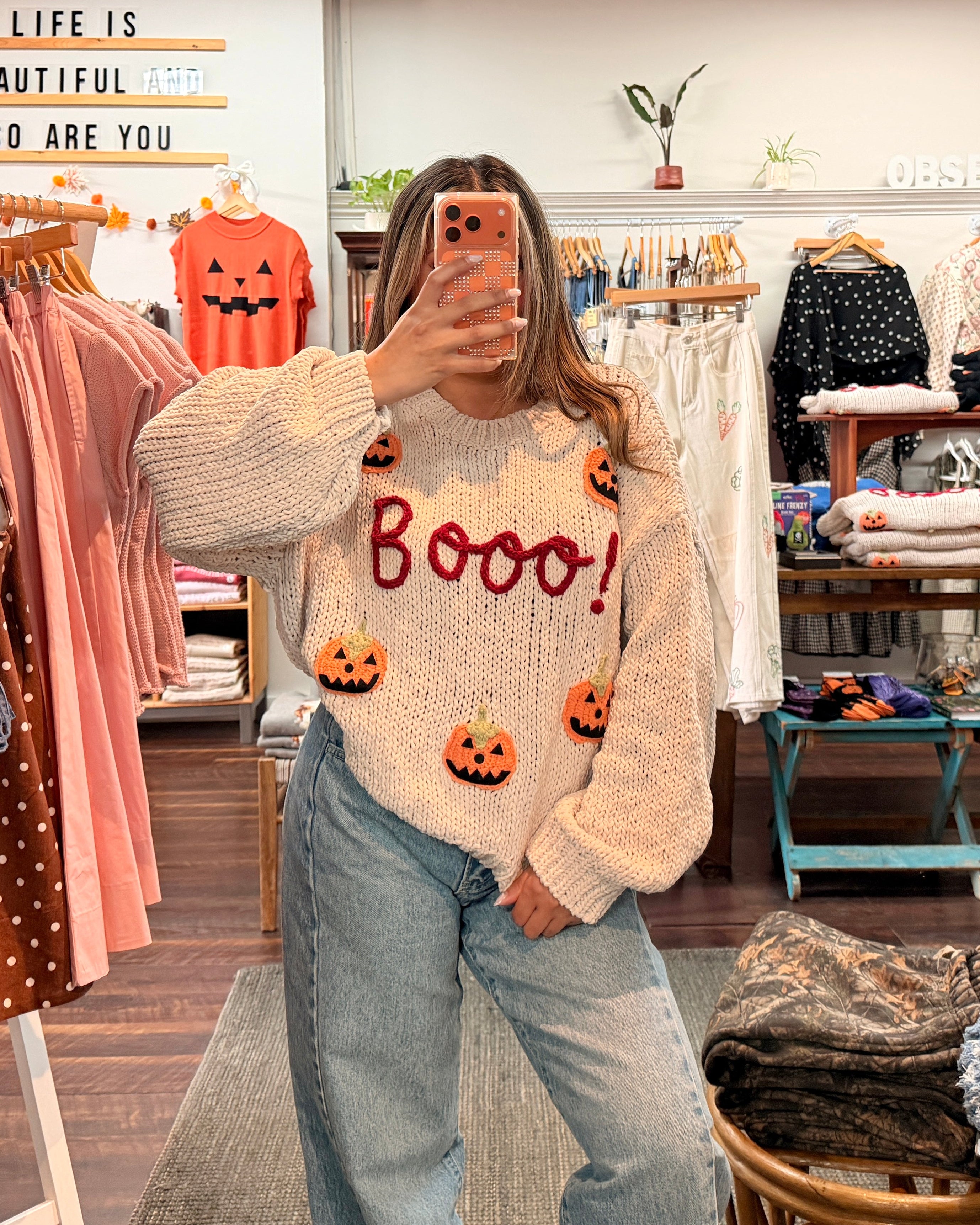 A woman takes a mirror selfie in a clothing boutique wearing a cream-colored knit sweater with 'Booo!' in red and small orange pumpkin graphics, paired with light-wash wide-leg jeans.
