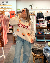 A woman takes a mirror selfie in a clothing boutique wearing a cream-colored fuzzy sweater with 'Boooo!' and pumpkin details, paired with light-wash blue jeans.