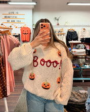 A woman takes a mirror selfie in a clothing boutique wearing a cream-colored fuzzy knit sweater with 'Boooo!' in red and orange pumpkin graphics, paired with blue jeans.