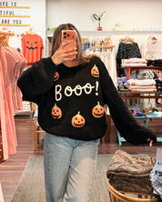 A woman takes a mirror selfie in a clothing boutique wearing a black fuzzy sweater with orange jack-o'-lantern graphics and the word 'Boooo!' in white. She is wearing light-wash blue jeans.