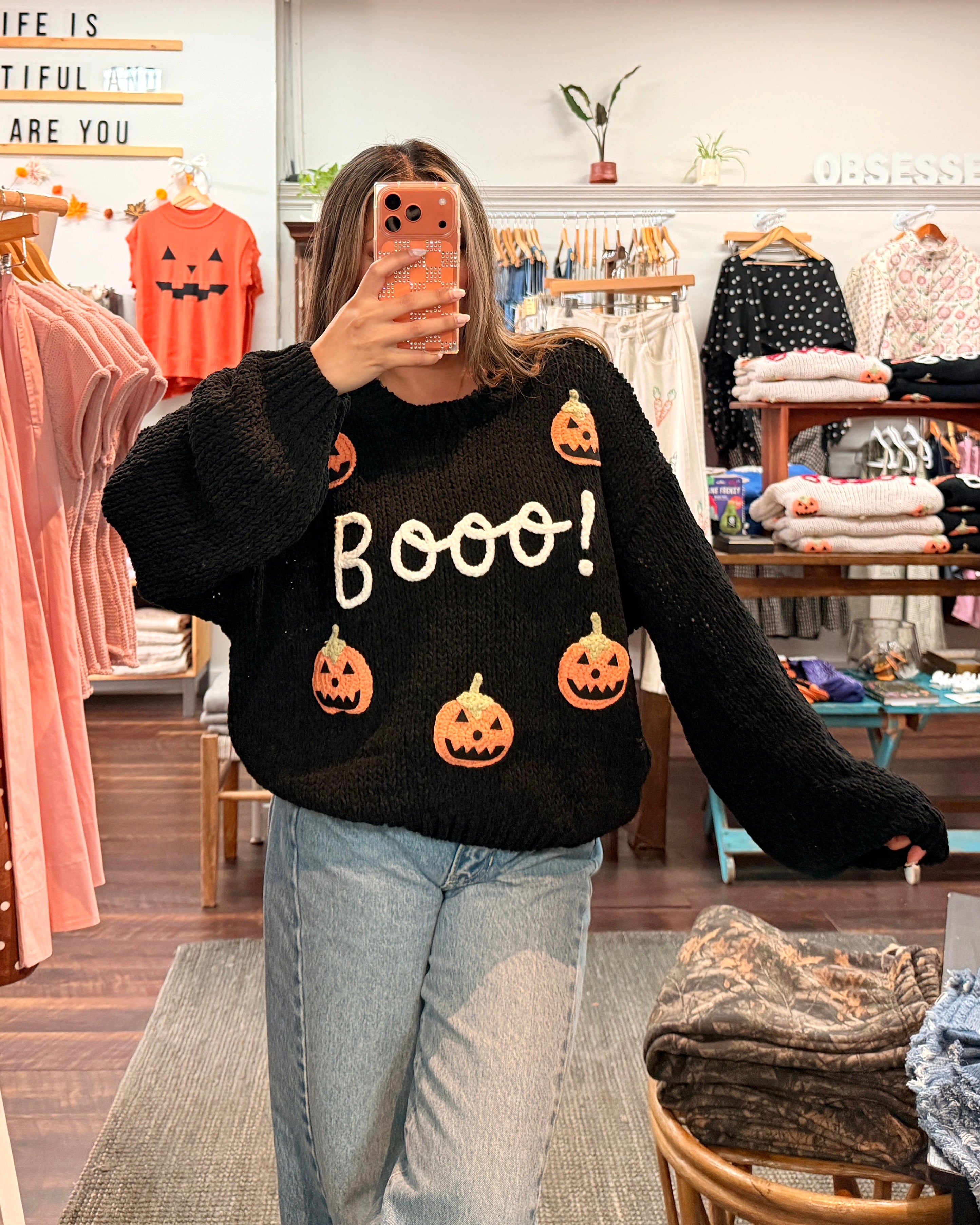 A woman takes a mirror selfie in a clothing boutique wearing a black fuzzy sweater with orange jack-o'-lantern graphics and the word 'Boooo!' in white. She is wearing light-wash blue jeans.