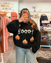 A woman takes a mirror selfie in a clothing boutique wearing a black crewneck sweater with 'Boooo!' in white text and orange pumpkin graphics, paired with light blue jeans.