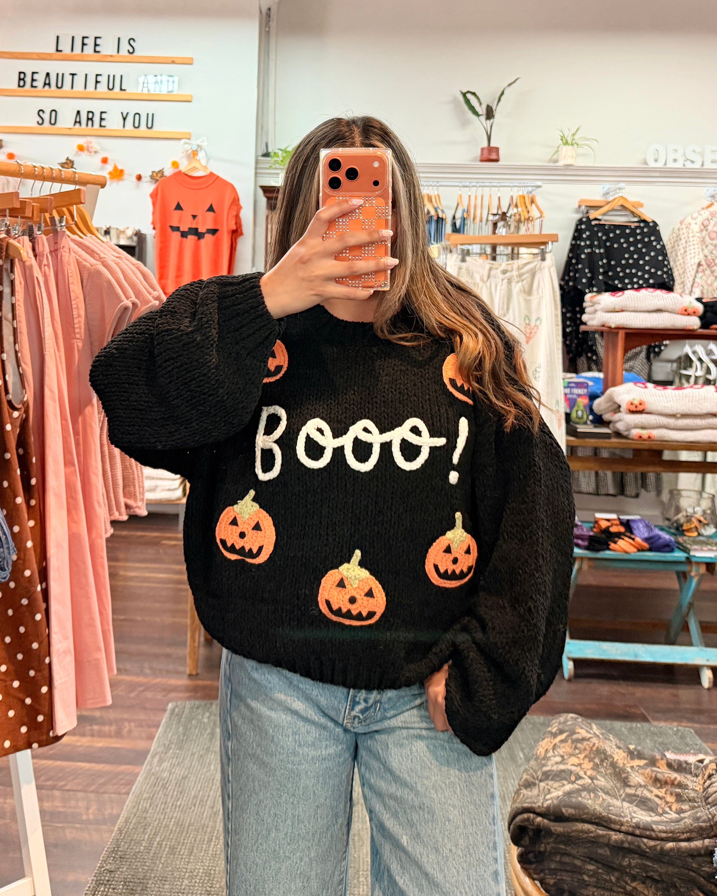 A woman takes a mirror selfie in a clothing boutique wearing a black crewneck sweater with 'Boooo!' in white text and orange pumpkin graphics, paired with light blue jeans.