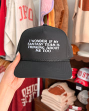 A hand holds a black trucker-style cap with white embroidered text reading 'I WONDERS IF MY FANTASY TEAM IS THINKING ABOUT ME TOO' in a retail clothing store setting with shirts and accessories in the background.