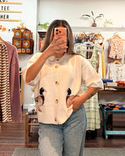 A woman takes a mirror selfie in a clothing boutique, wearing a cream-colored short-sleeve button-up shirt with knit duck detailing