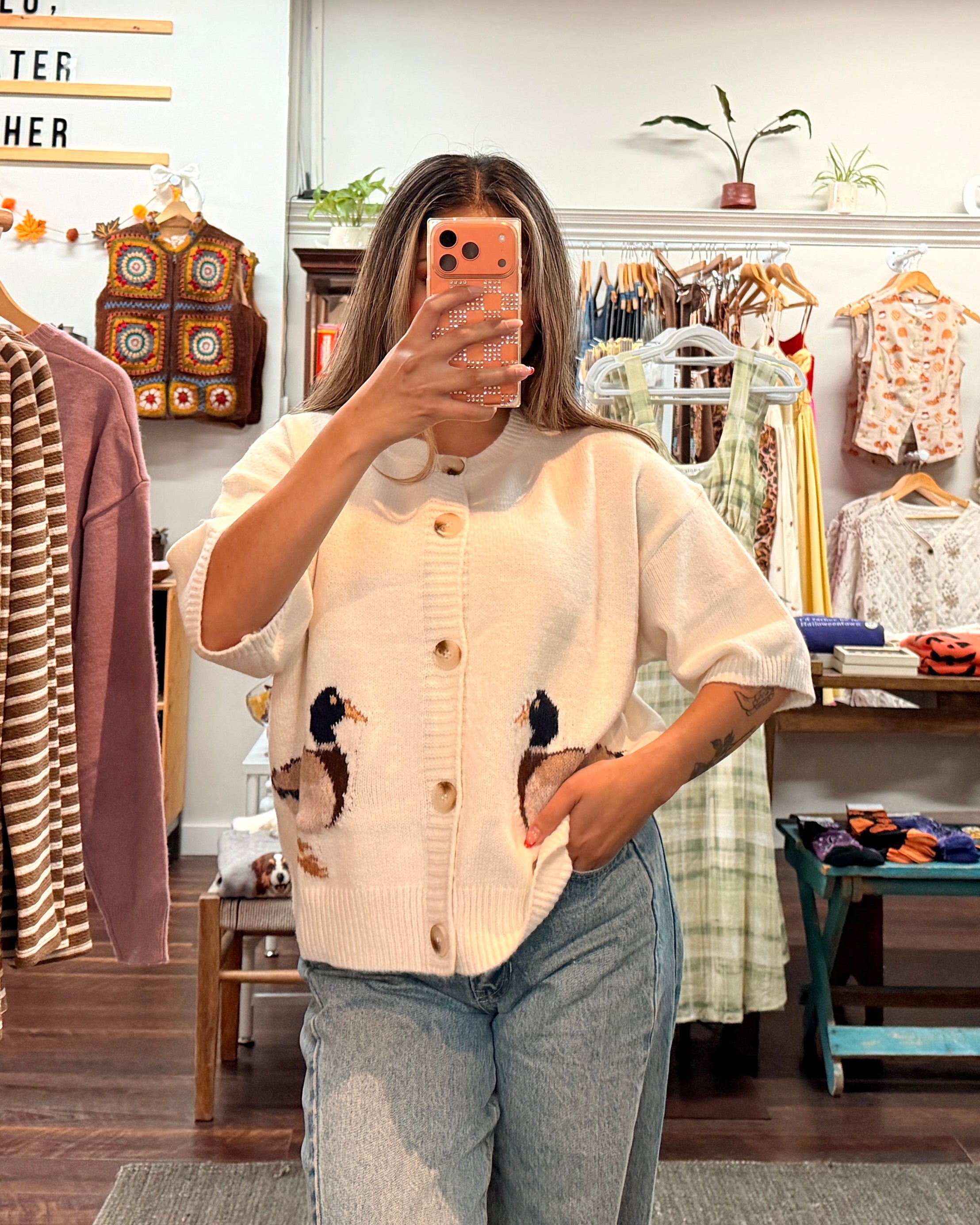 A woman takes a mirror selfie in a clothing boutique, wearing a cream-colored short-sleeve button-up shirt with knit duck detailing