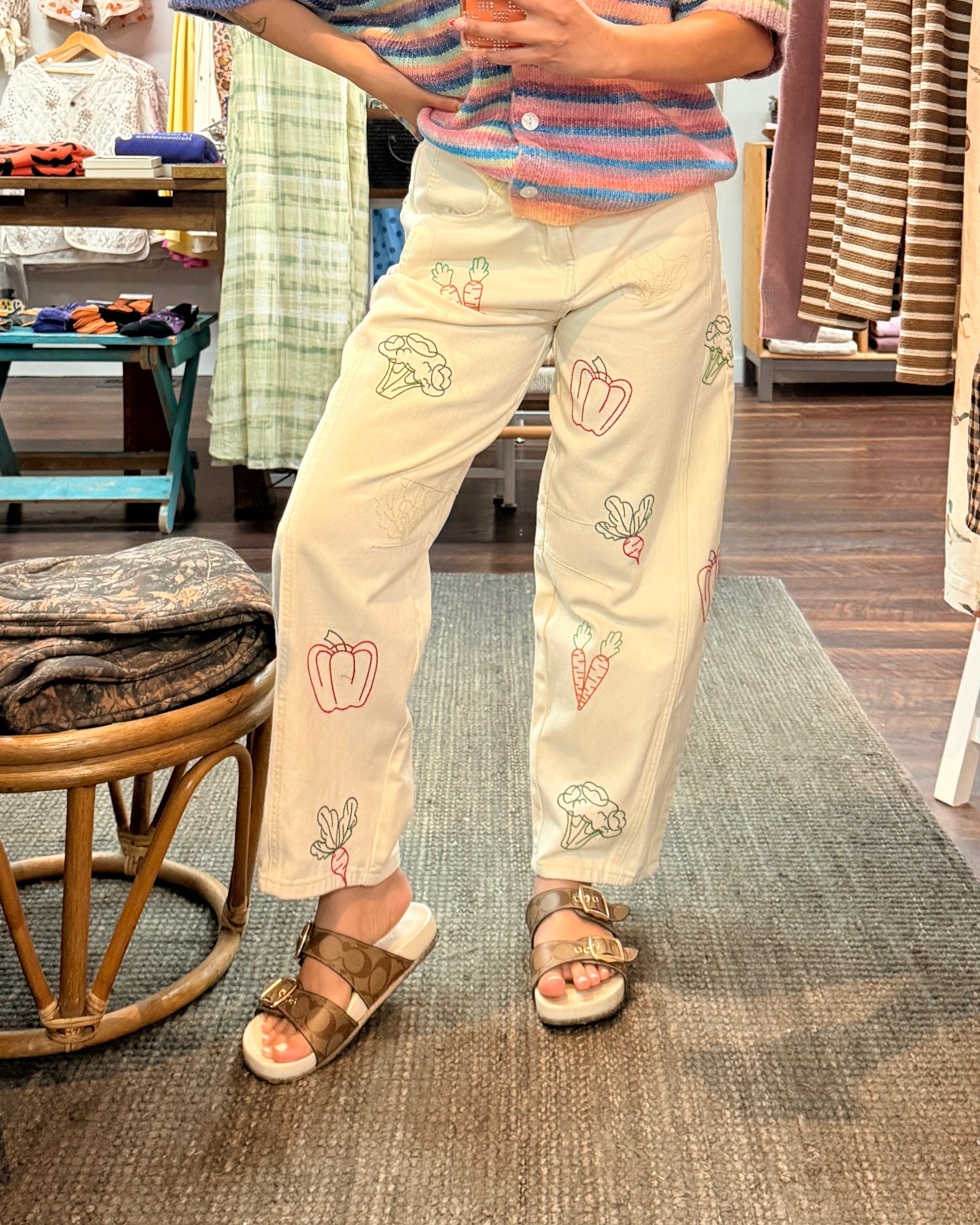A woman takes a mirror selfie in a clothing boutique, wearing a pastel rainbow striped t-shirt and cream-colored pants with colorful embroidered vegetable details.
