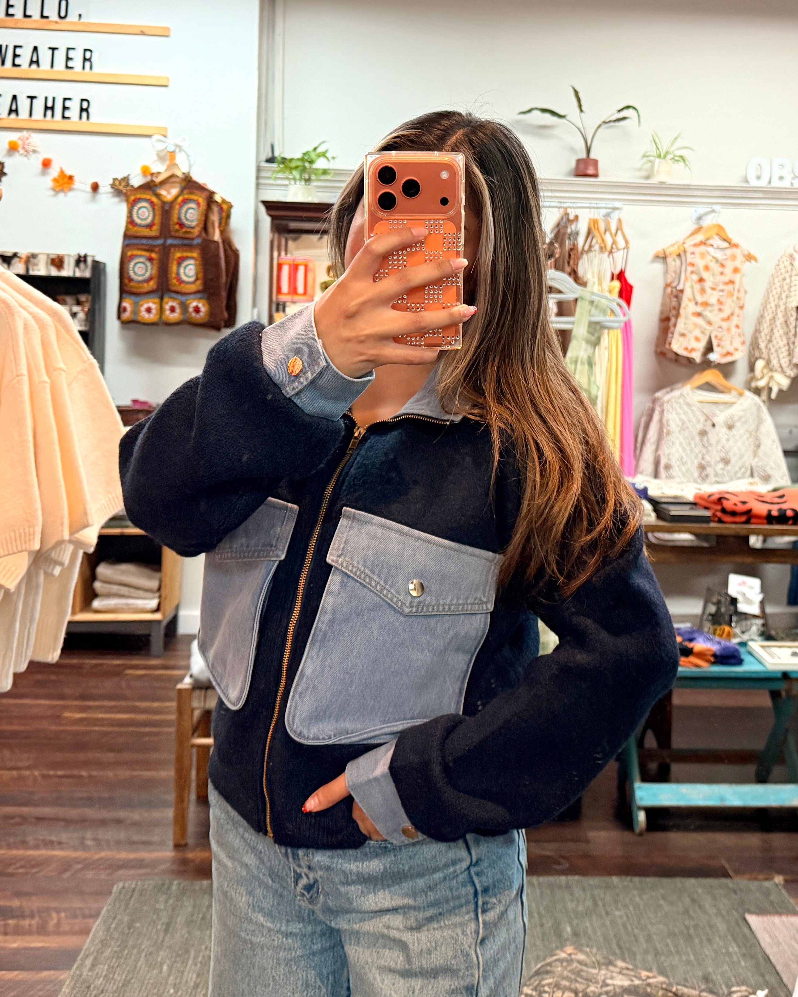 A person taking a mirror selfie in a clothing boutique, wearing a color-blocked zip-up jacket with navy, light blue, and gray panels, paired with light-wash blue jeans.
