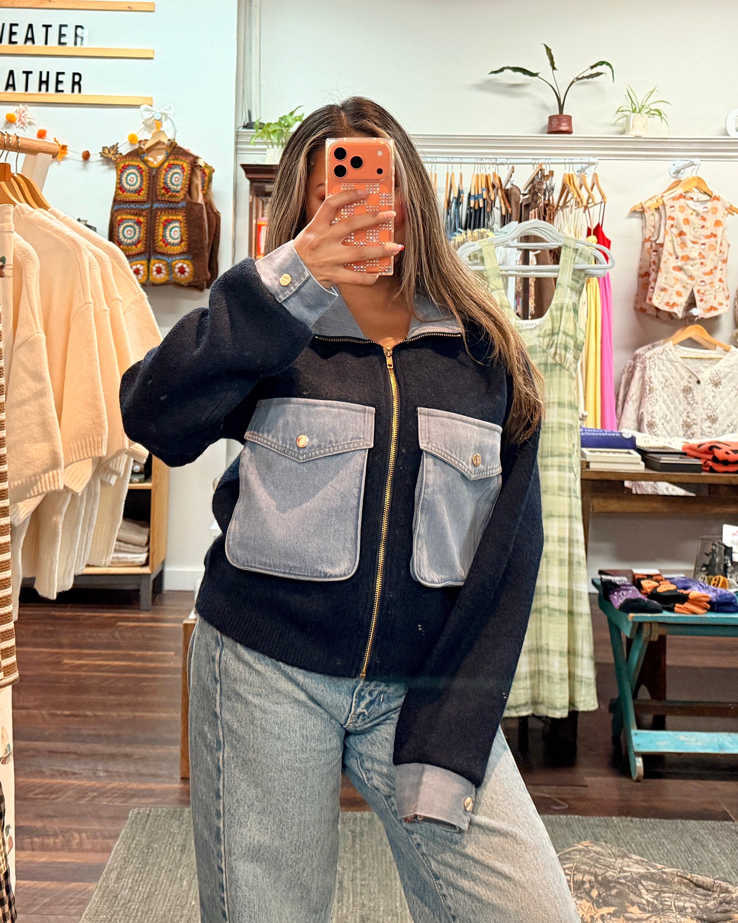 A woman takes a mirror selfie in a clothing boutique wearing a navy blue zip-up jacket with light blue denim pockets and light-wash blue jeans.