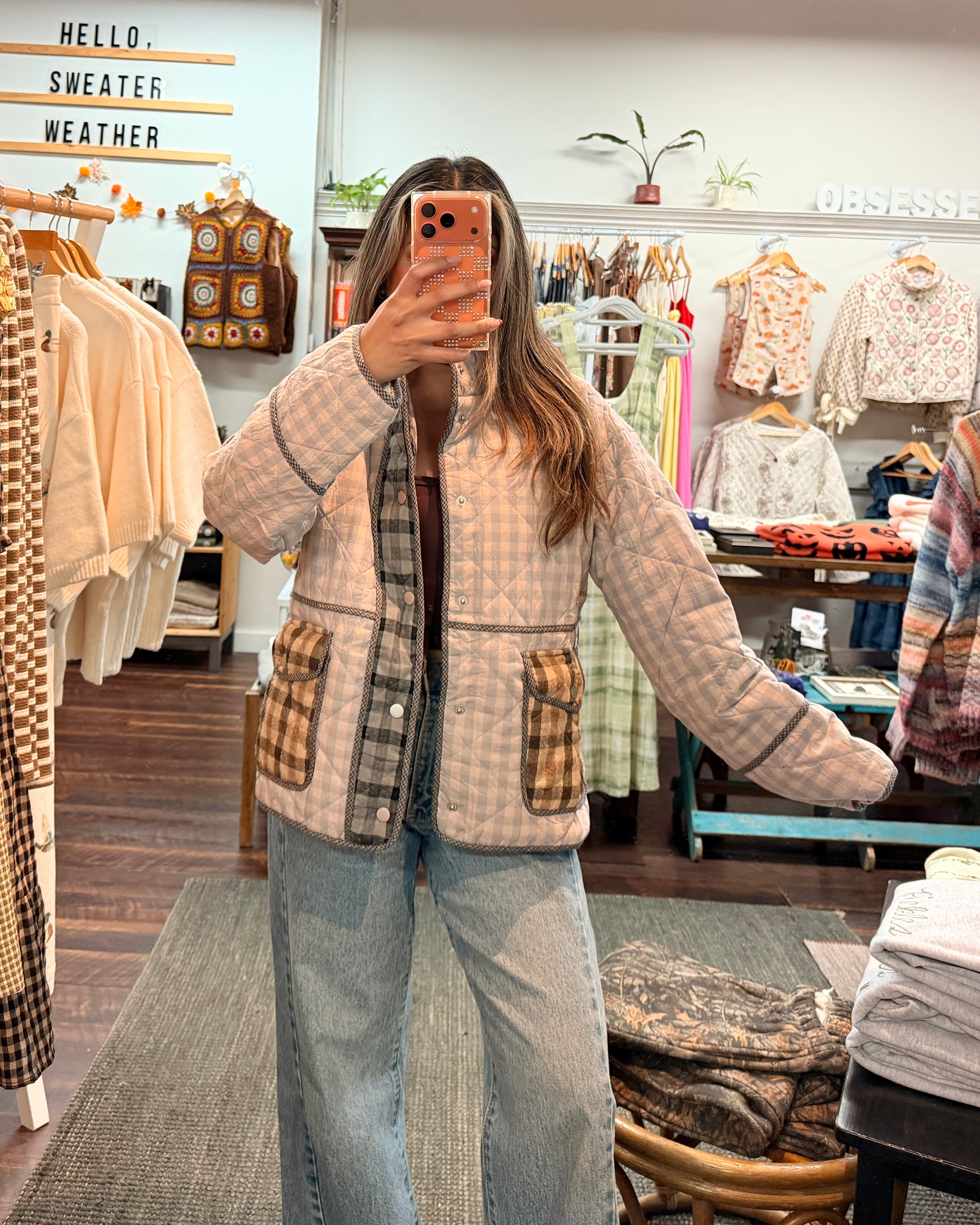 A woman takes a mirror selfie in a clothing boutique wearing a beige and brown plaid zip-up jacket with patch pockets and light blue wide-leg jeans. The boutique is filled with racks of colorful clothing.