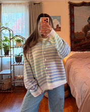 A woman takes a mirror selfie in a bedroom, wearing a pastel striped button-up cardigan with blue jeans. The room features a wooden floor, a bed with pink bedding, and a large tapestry on the wall.