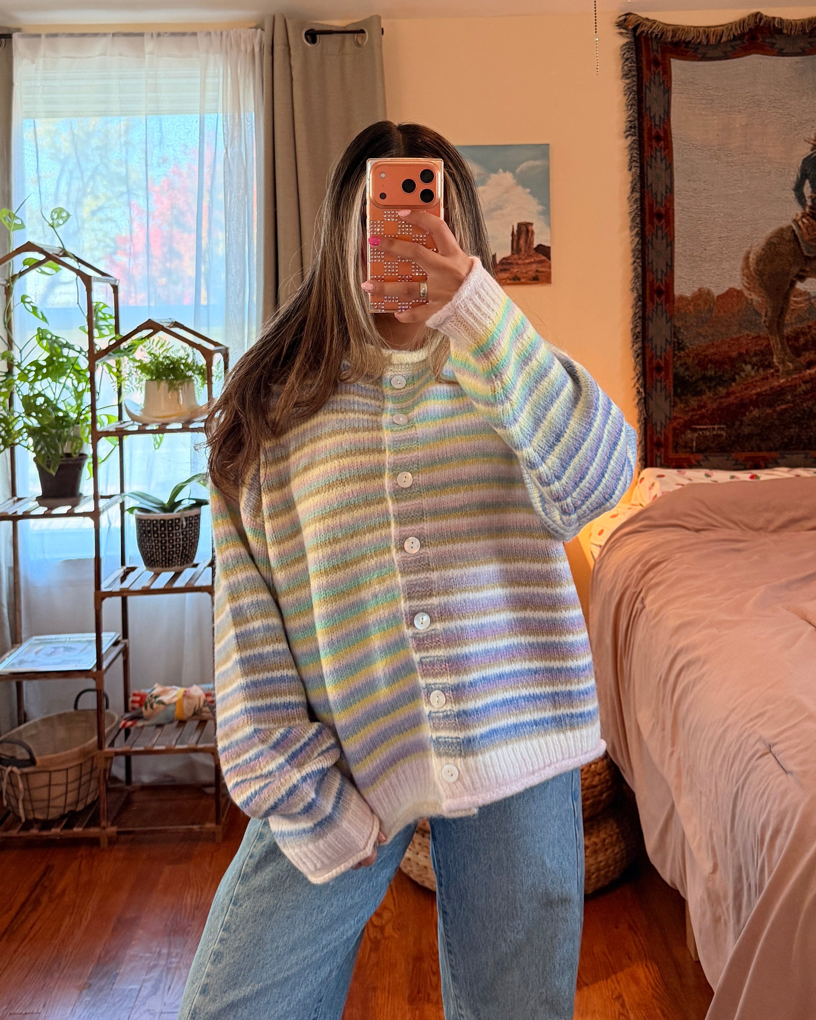 A woman takes a mirror selfie in a bedroom, wearing a pastel striped button-up cardigan with blue jeans. The room features a wooden floor, a bed with pink bedding, and a large tapestry on the wall.
