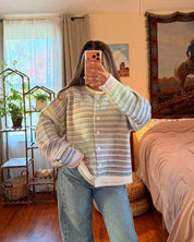 A person takes a mirror selfie in a bedroom, wearing a pastel striped knit cardigan with light blue jeans. The room features a wooden floor, a bed with beige bedding, and a large framed western-style tapestry.