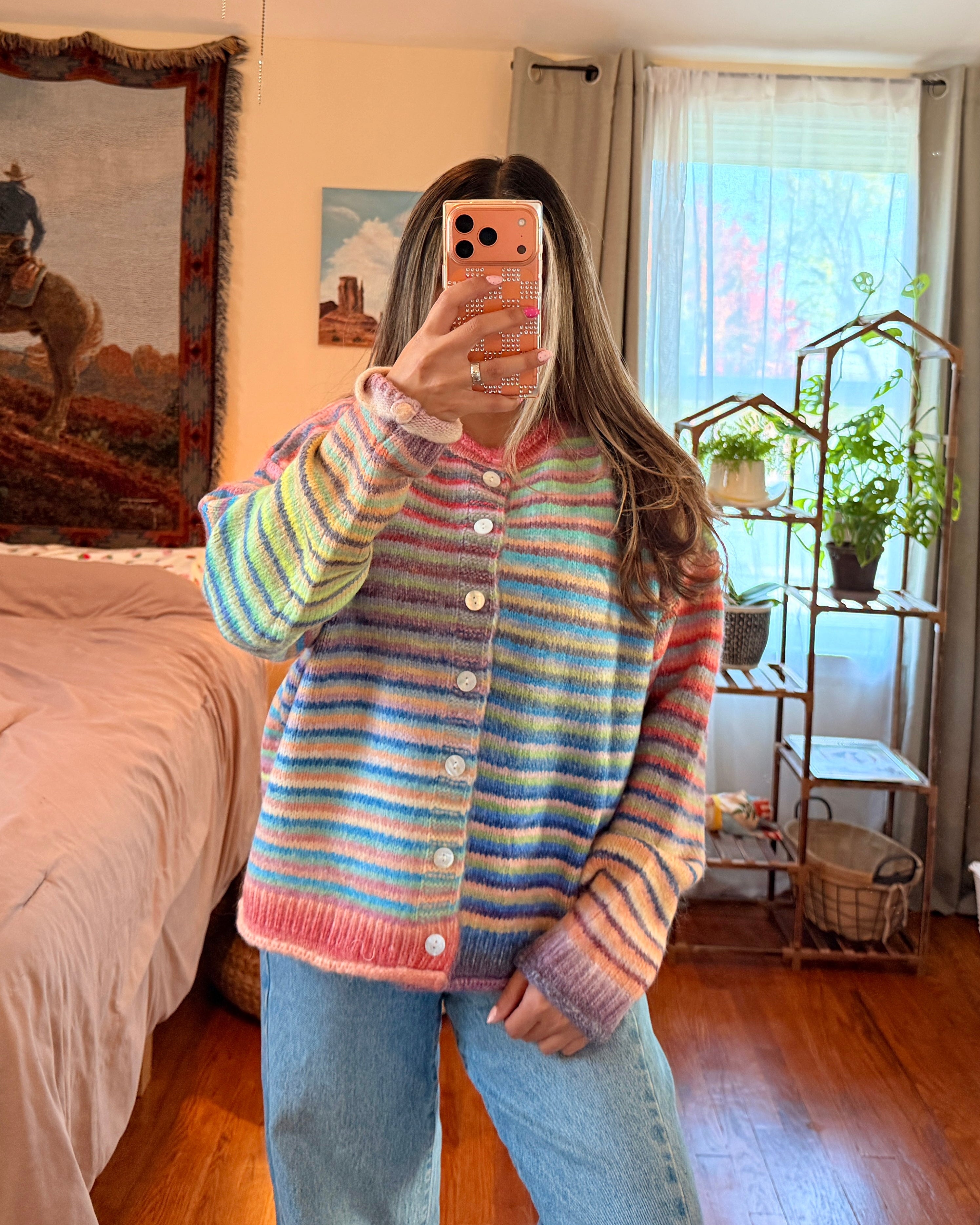 A woman takes a mirror selfie in a bedroom, wearing a pastel striped button-up cardigan and light blue jeans. The cardigan features horizontal stripes of pink, blue, green, yellow, and white, with a pink ribbed trim and buttons.