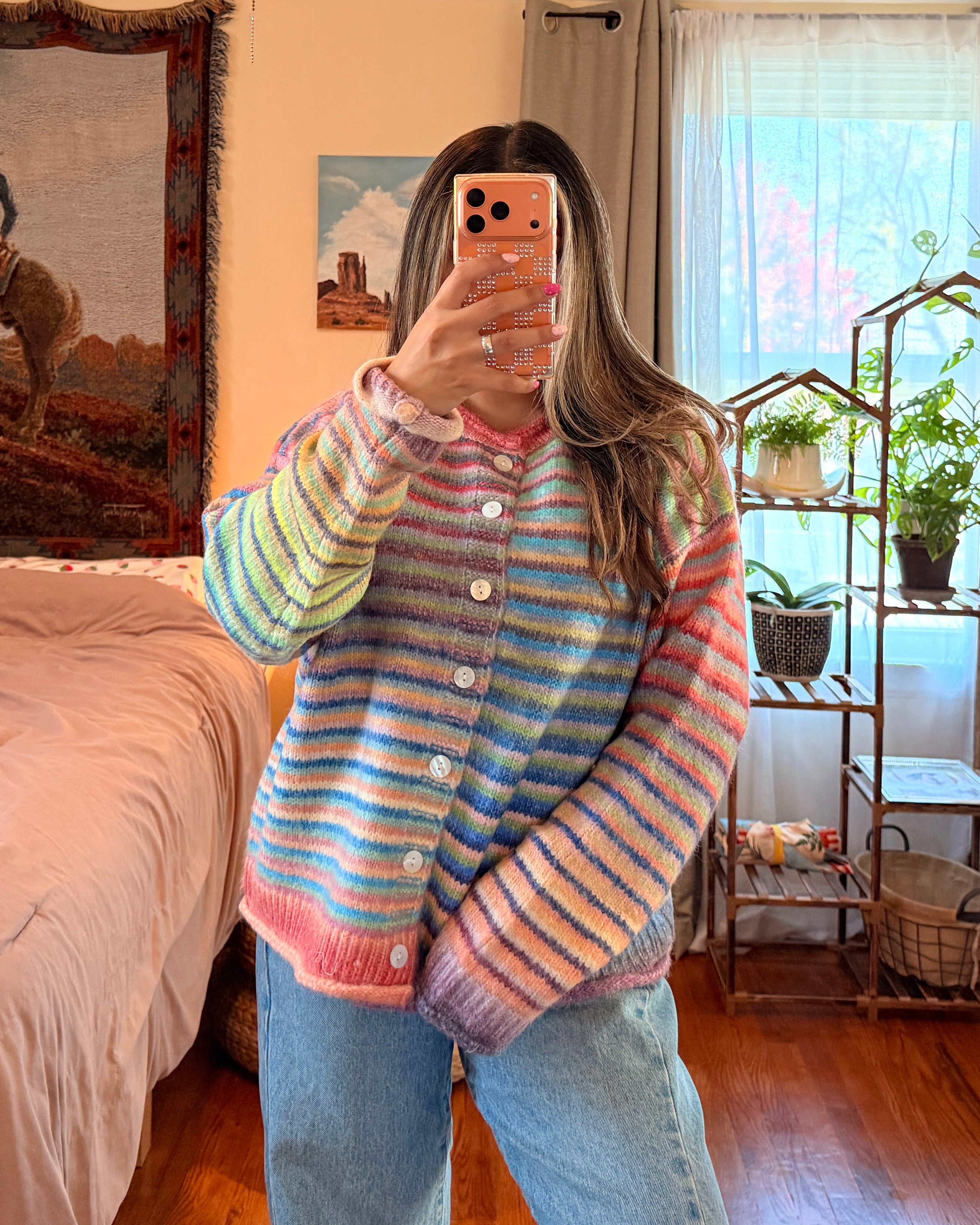 A person taking a mirror selfie in a bedroom, wearing a pastel rainbow striped button-up cardigan sweater and light blue jeans.