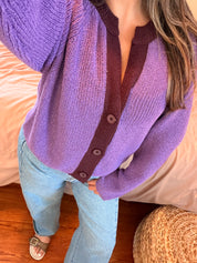 A person wearing a purple and dark burgundy knit cardigan with buttons, paired with light blue jeans, standing in a bedroom setting.