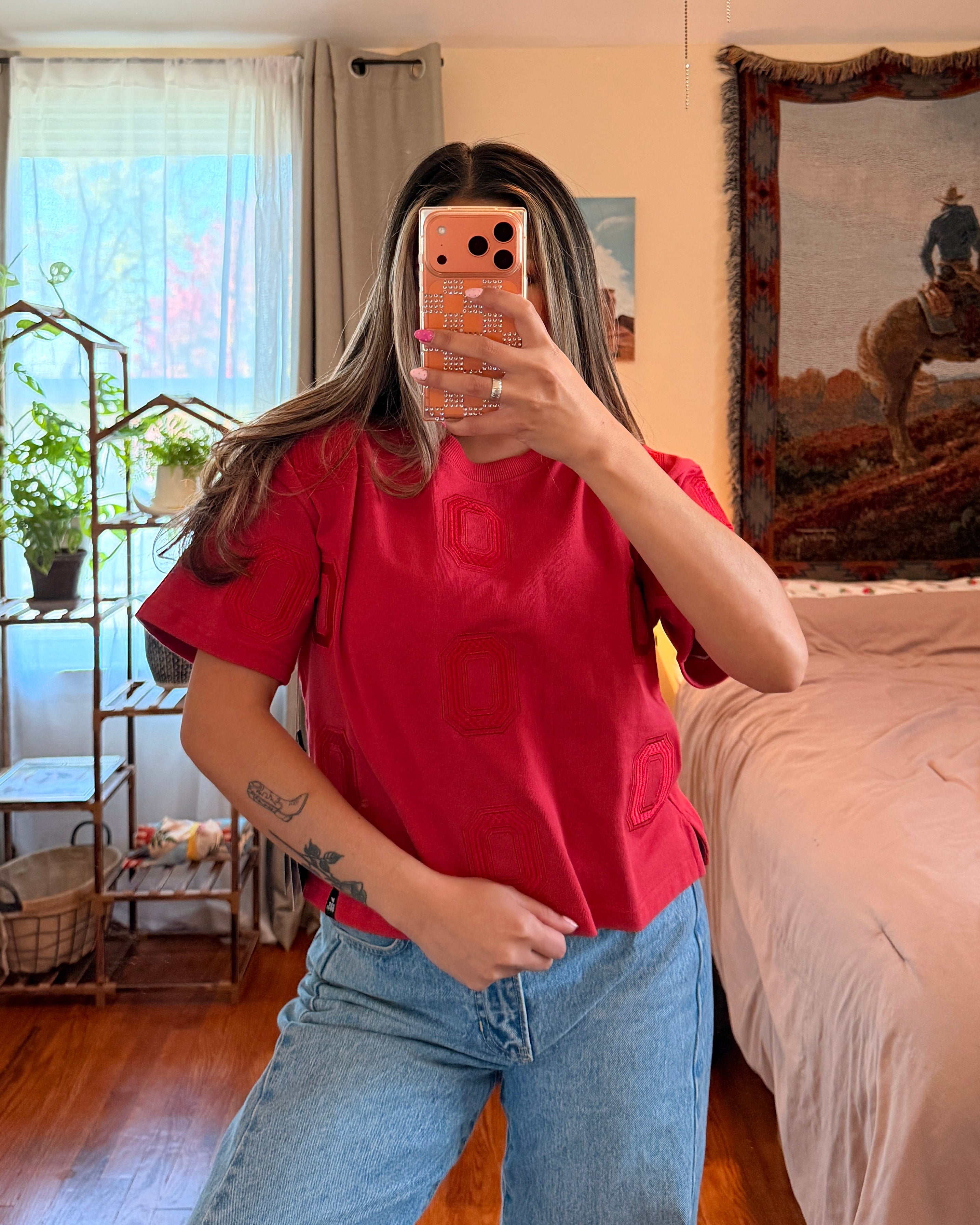 A woman takes a mirror selfie in a bedroom, wearing a bright red short-sleeve t-shirt and light blue jeans. The room features a wooden floor, a bed with pink bedding, and a western-themed tapestry on the wall.