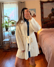 A woman takes a mirror selfie in a bedroom wearing a cream-colored knit dress and yellow boots.