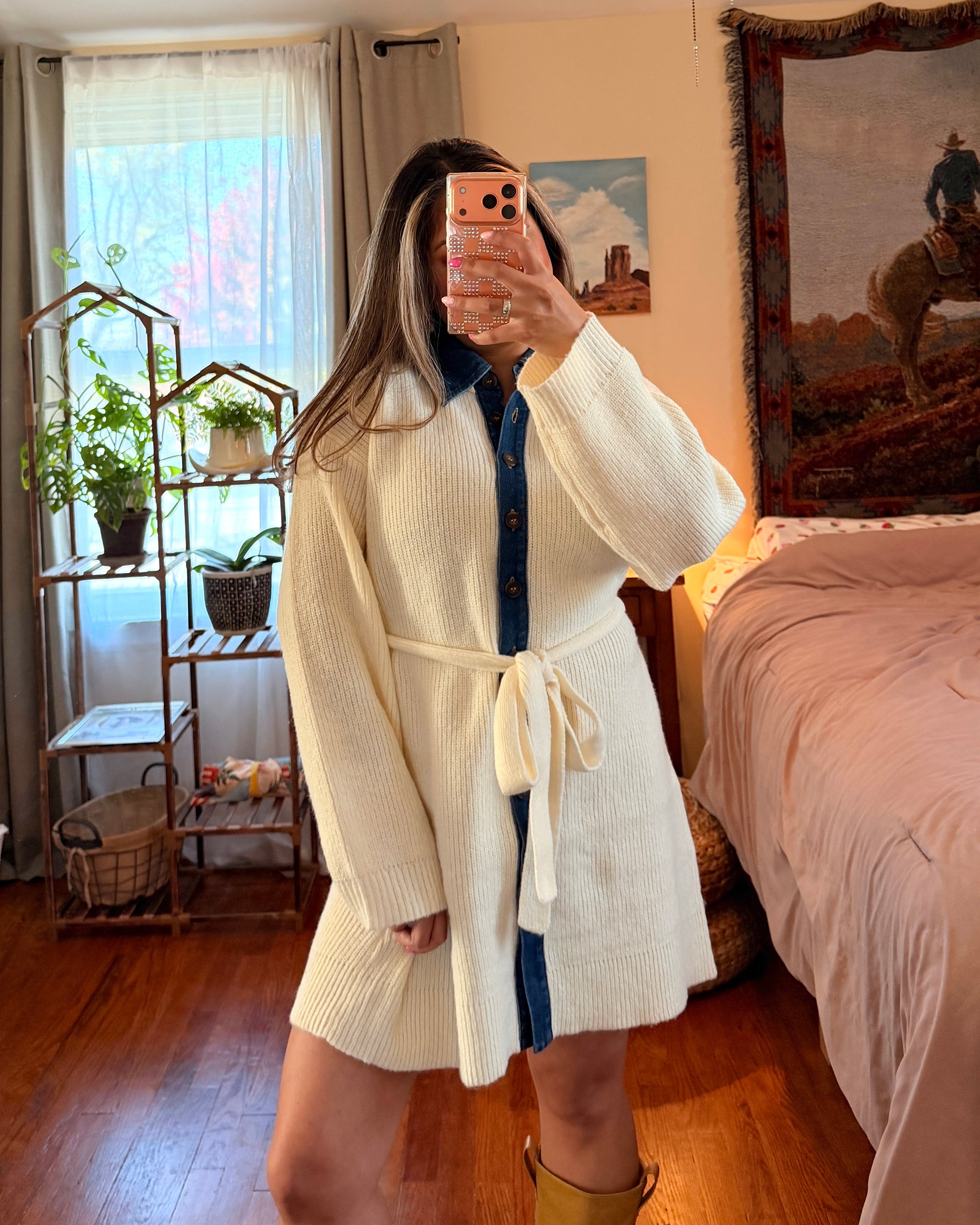 A woman takes a mirror selfie in a bedroom wearing a cream-colored knit dress and yellow boots.