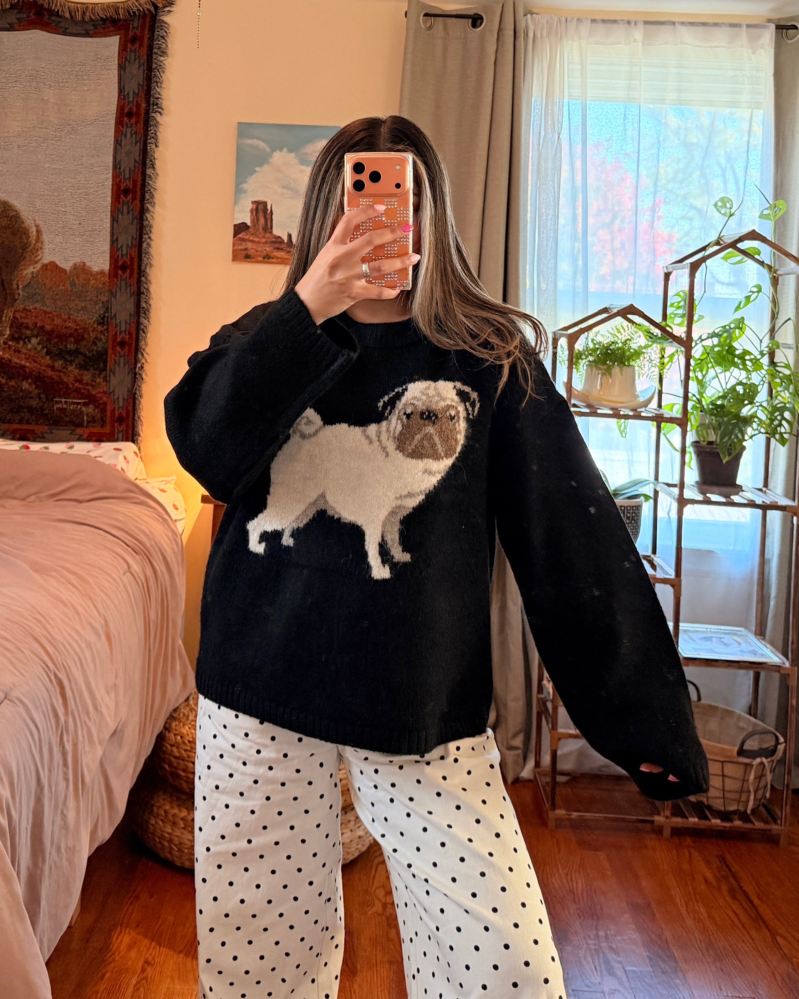 A person taking a mirror selfie in a bedroom, wearing a black long-sleeve sweatshirt with a white pug graphic and white pants with black polka dots.