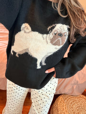 A close-up of a person wearing a black knit sweater with a large white and brown pug dog graphic across the front, paired with black polka dot pants, in a cozy indoor setting.
