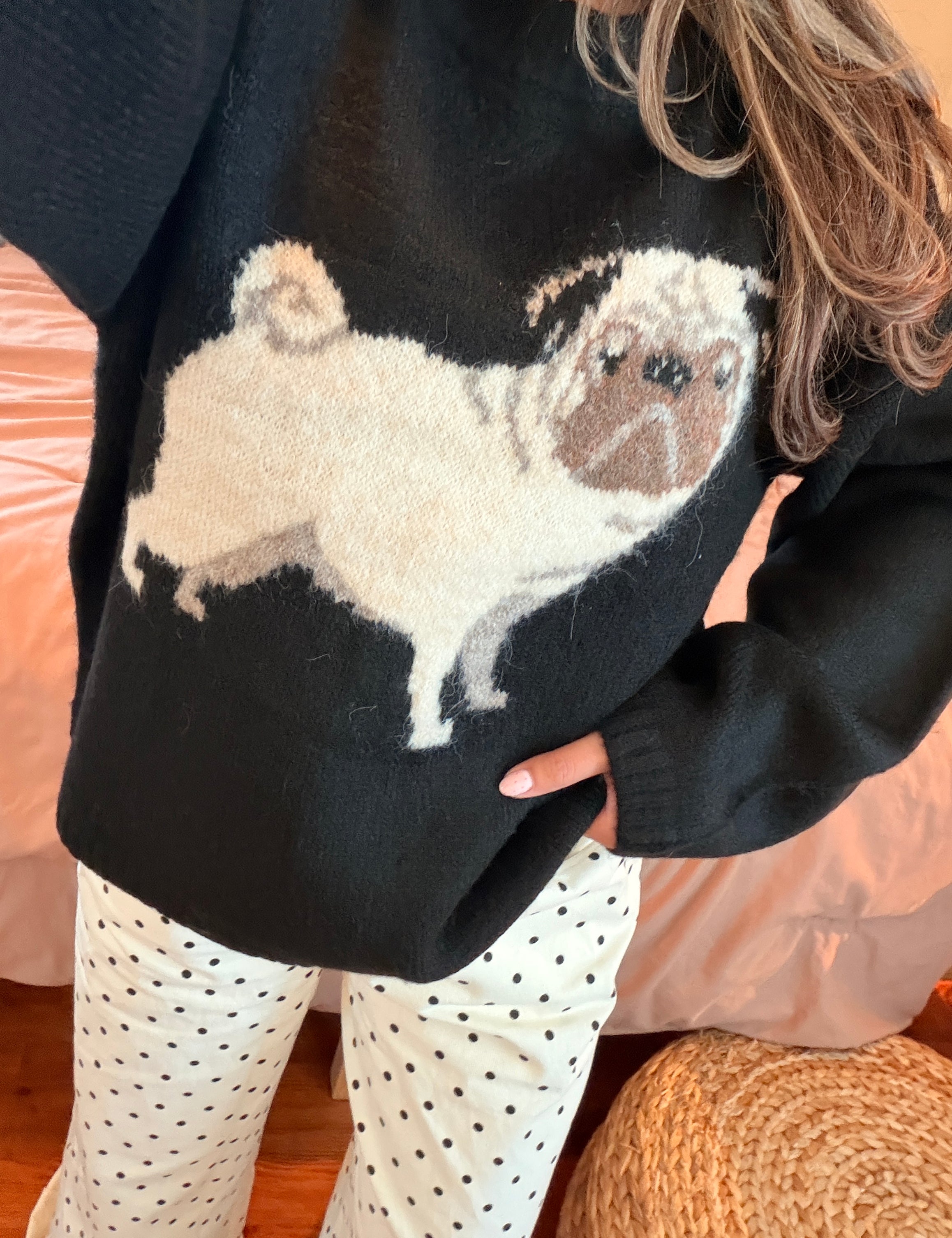 A close-up of a person wearing a black knit sweater with a large white and brown pug dog graphic across the front, paired with black polka dot pants, in a cozy indoor setting.