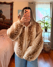 A person taking a mirror selfie in a bedroom, wearing a beige faux fur jacket with white spots resembling a deer print, paired with blue jeans.